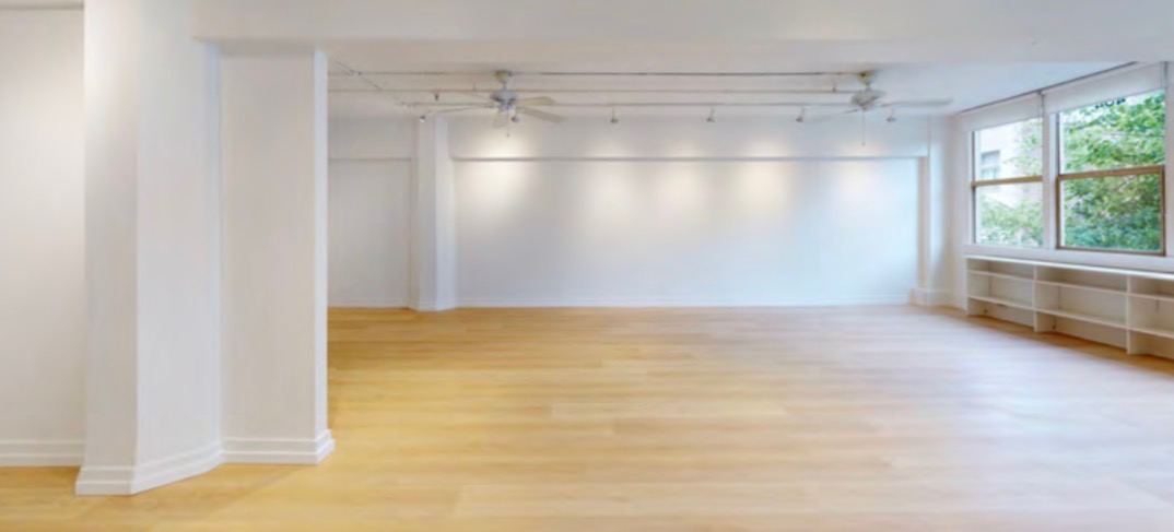 The open main studio: white walls and columns, pale natural wood floor, track lighting, ceiling fans, and windows with greenery along the right wall.