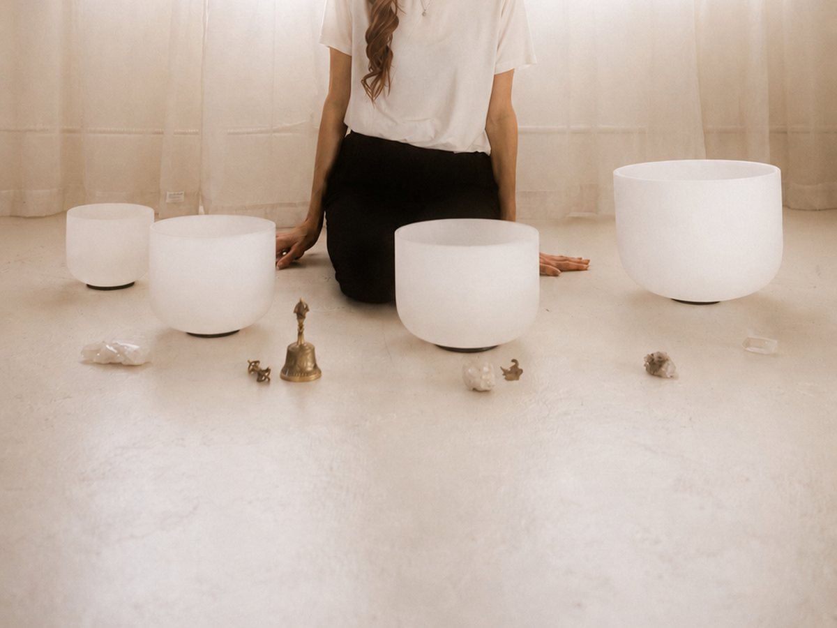 A practitioner kneeling behind white crystal singing bowls, a small bell and crystals on a pale floor.